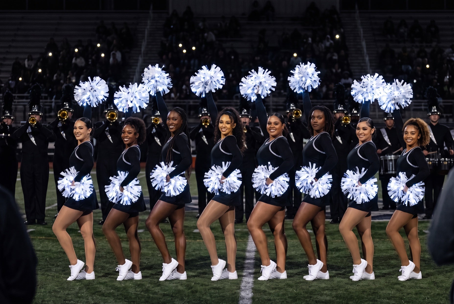 Band dance line: eight dancers in black hold white LED pom poms in front of a marching band on a dark field