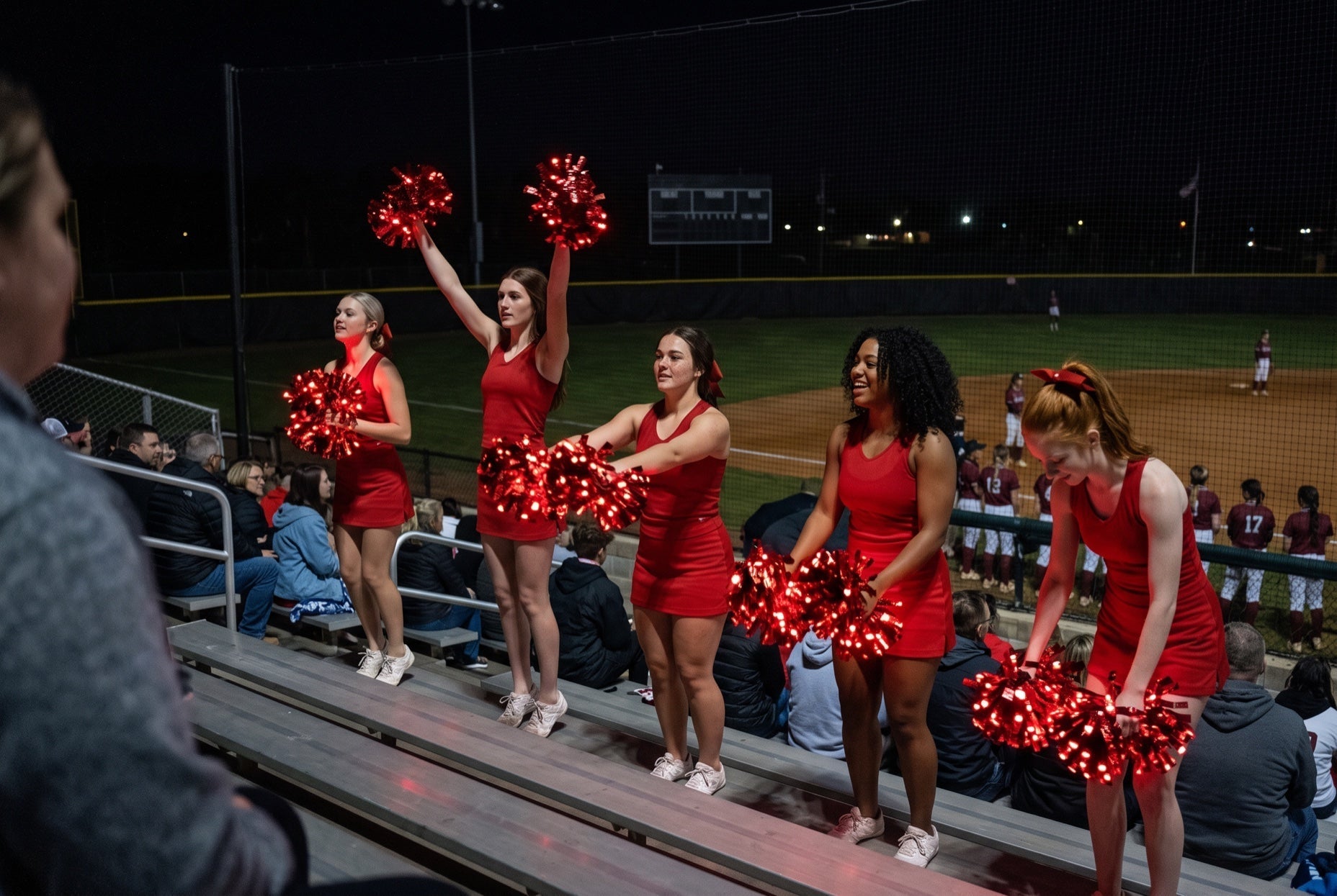 Light up LED pom poms glowing red on a high school cheer squad in the bleachers at a night softball game