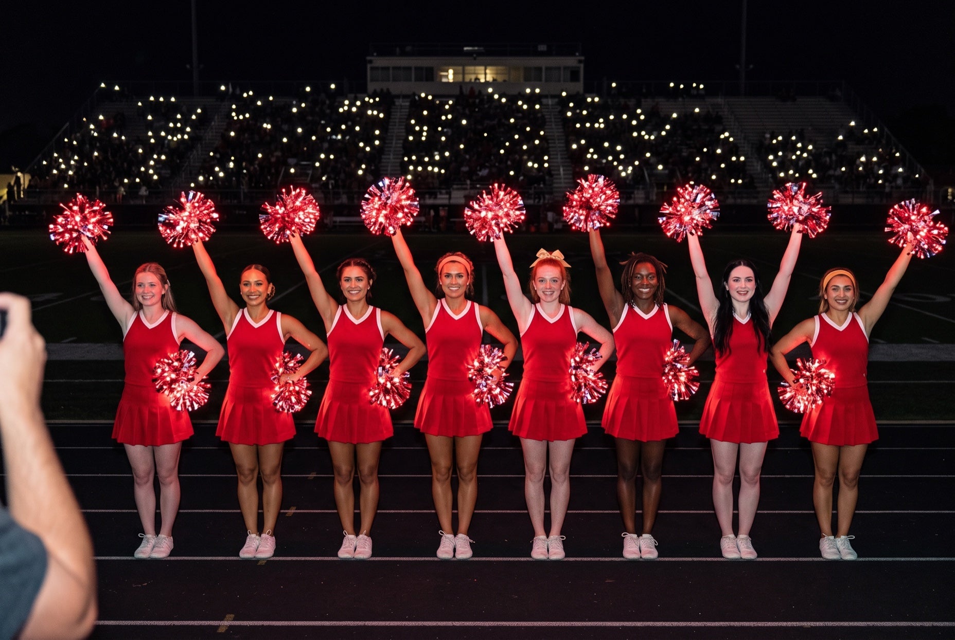 Cheer recruitment game night: college cheerleaders in red raise red LED pom poms before dark stands full of phone lights