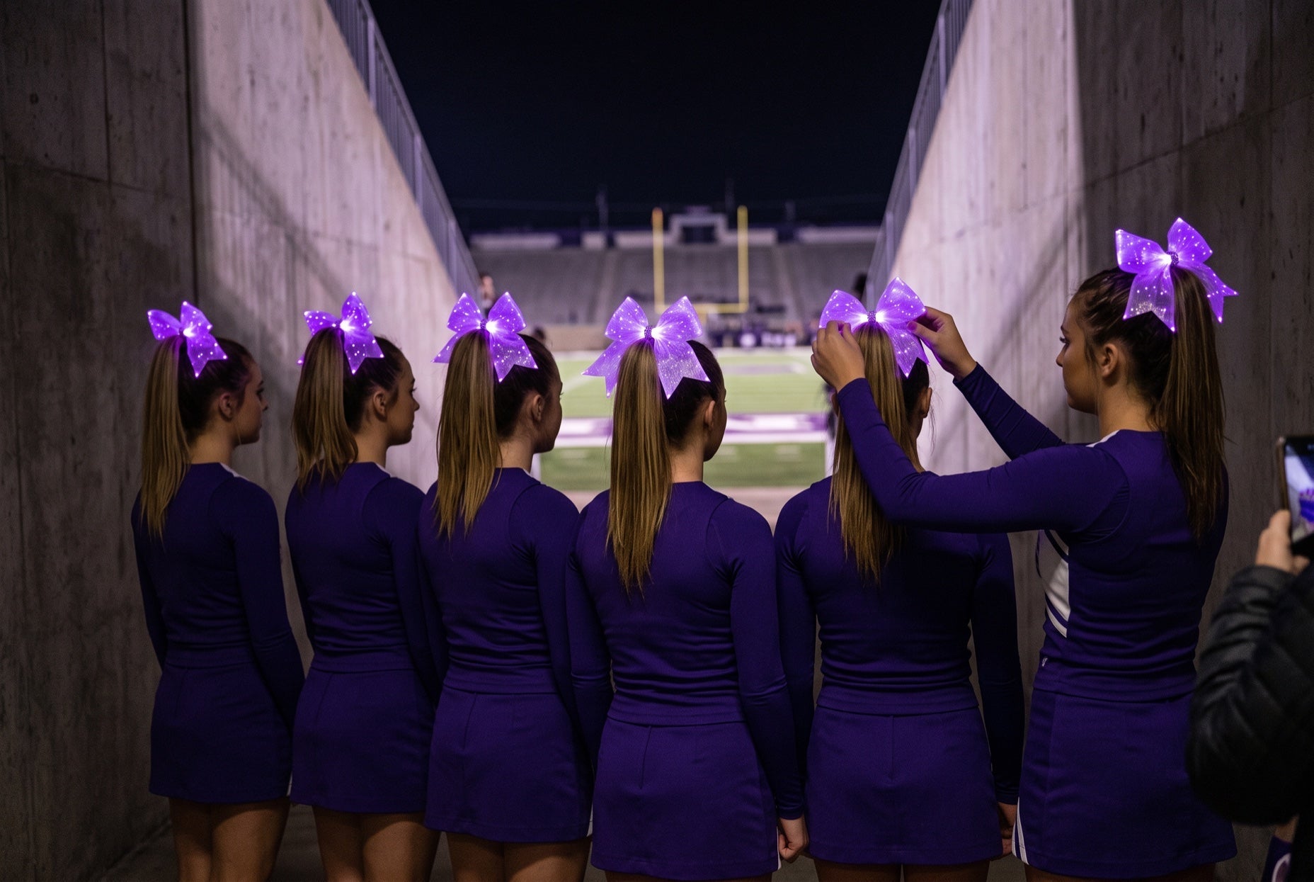 Cheer bow placement: a captain straightening a glowing purple LED cheer bow at the crown of a teammate's high ponytail