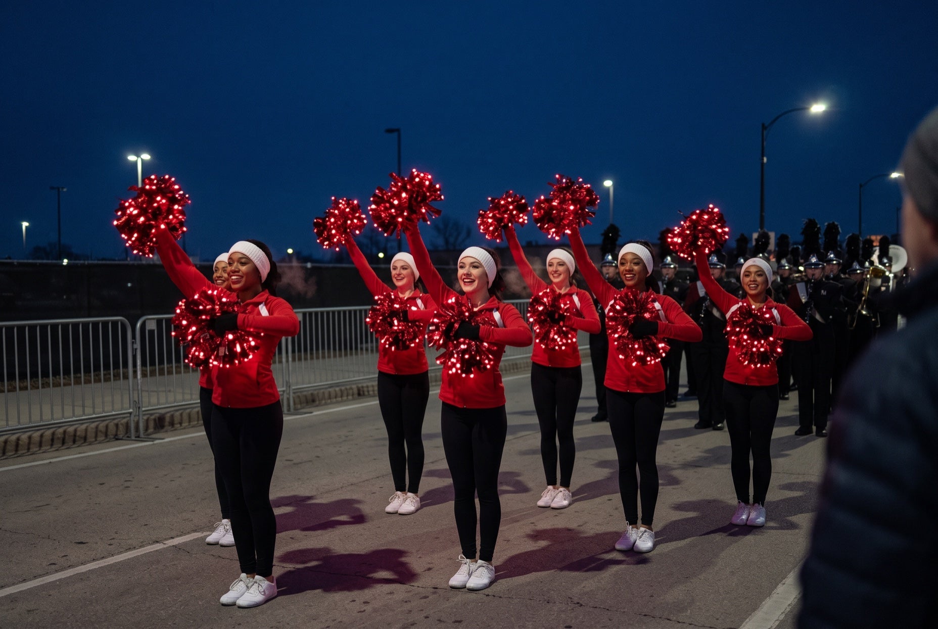 Cheer squad with glowing red LED light-up pom poms at a Thanksgiving morning parade in the pre-dawn dark, band behind