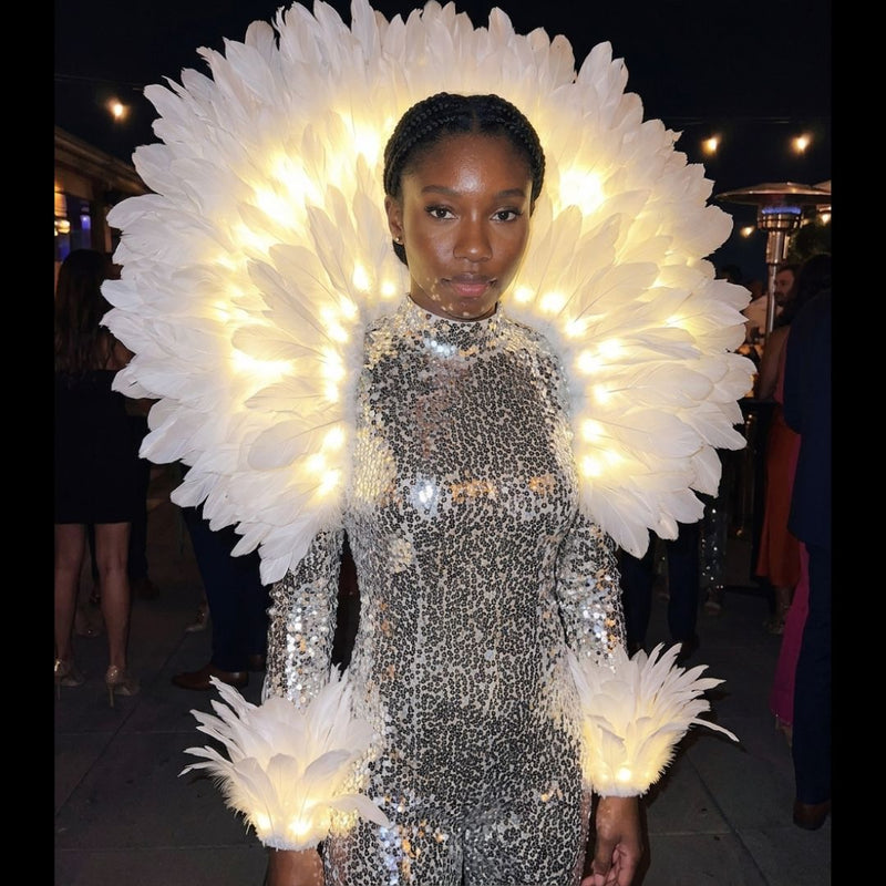 Woman wearing a glowing LED feather aura back piece with matching light-up feather arm cuffs over a silver sequin outfit at an elegant evening event — an illuminated feather halo and elegant light-up accessory for performers, showgirls, galas, festivals, burlesque, circus and stage and event entertainers.