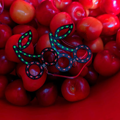 Bright red LED cherry stickers displayed on actual cherries - Creative product showcase of illuminated fruit-shaped facial jewelry for food-themed parties, summer festivals, and fruit fashion trends