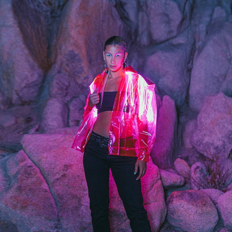 A model in a glowing translucent pink LED fringe jacket posing against boulders at night, a custom light up fringe jacket and led jacket creating eye catching movement, glowing stagewear for go-go dancers, drag and burlesque performers at music festivals, EDM concerts and nightclub parties, Neon Cowboys