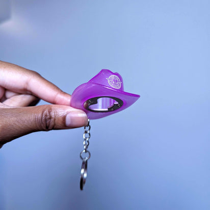 Hand holding purple cowboy hat keychain bottle opener demonstrating size and usability at concerts and nighttime gatherings