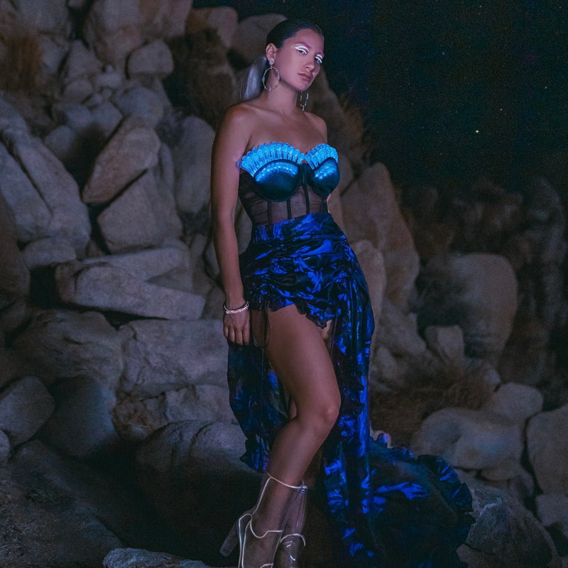 Model in a Neon Cowboys high low floral bustle skirt with a glowing blue LED bulb corset on rocks, illuminated light up festival stagewear for go-go dancers, burlesque performers and showgirls, dramatic silhouette for music festivals, theatrical performances and nightlife events, Neon Cowboys