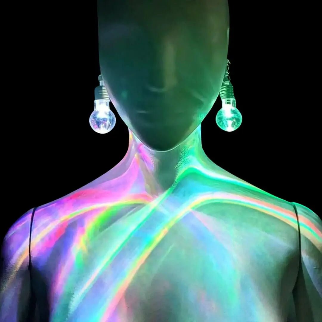 Woman wearing color-changing LED bulb earrings against dark background - Illuminated light bulb-shaped jewelry casting colorful reflections for music festivals, nightclub events, EDM concerts, and futuristic fashion looks