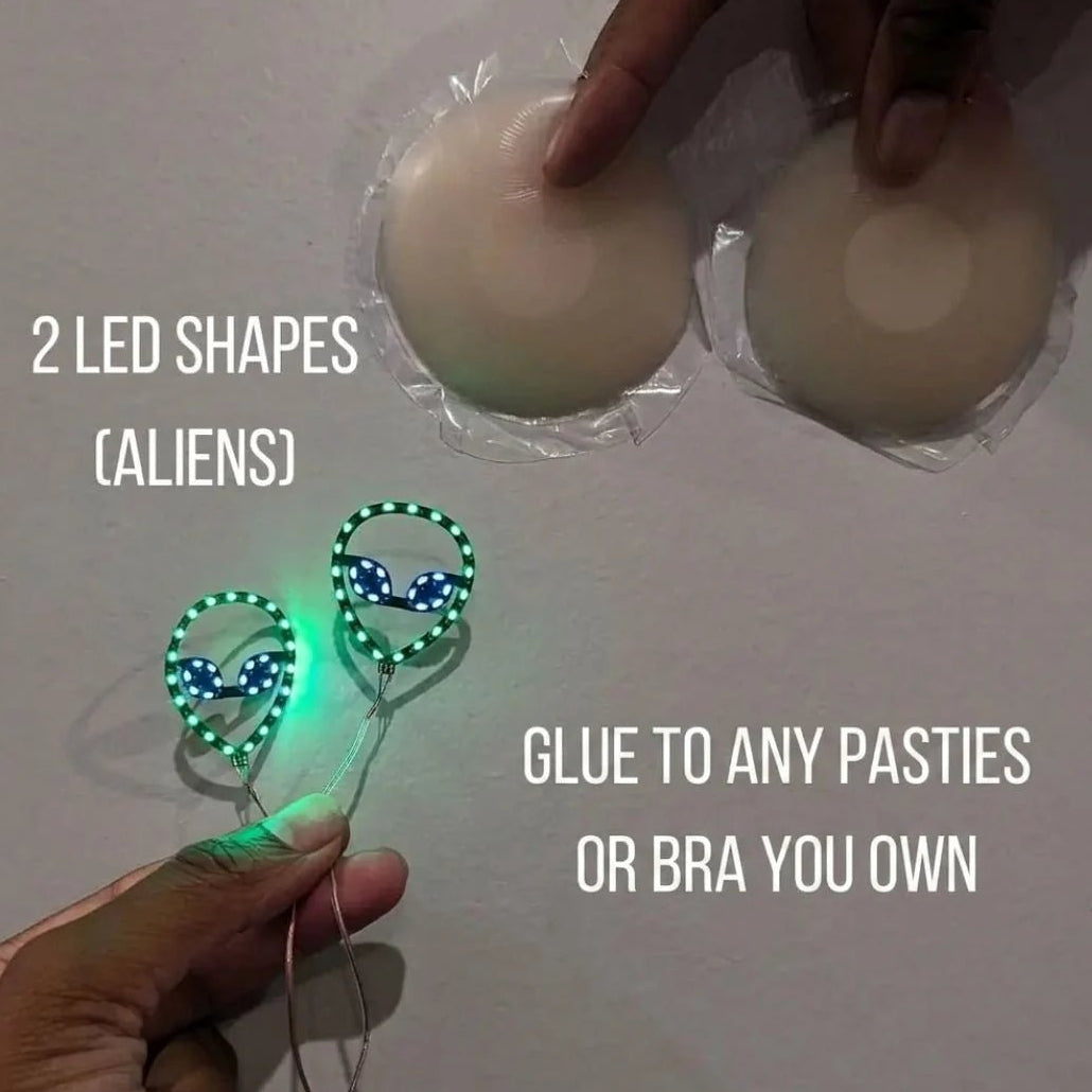 Two glowing green LED alien shapes next to silicone pasties — light-up alien nipple covers you glue onto any pasties or bra