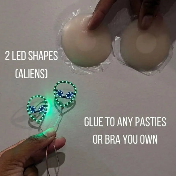 Two glowing green LED alien shapes next to silicone pasties — light-up alien nipple covers you glue onto any pasties or bra