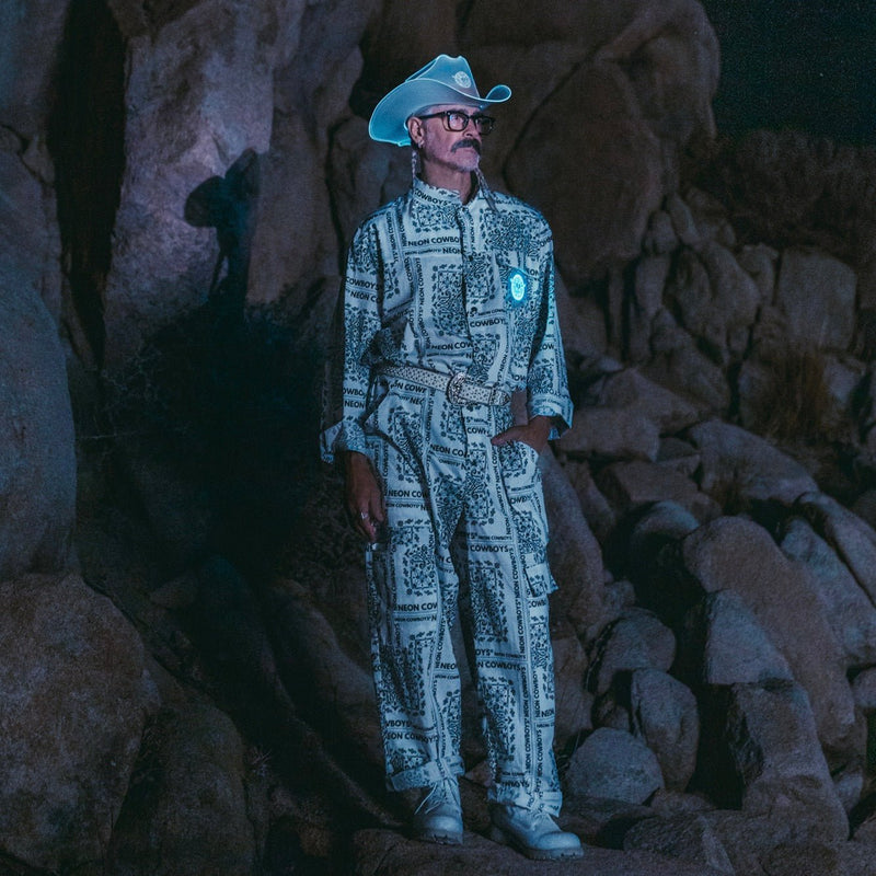 Man wears a Neon Cowboys light up bandana jumpsuit glowing blue with logo paisley print and a matching cowboy hat in the desert, led jumpsuit rave stagewear for go-go dancers and performers, perfect for music festivals, Stagecoach concert outfits and country western events, Neon Cowboys