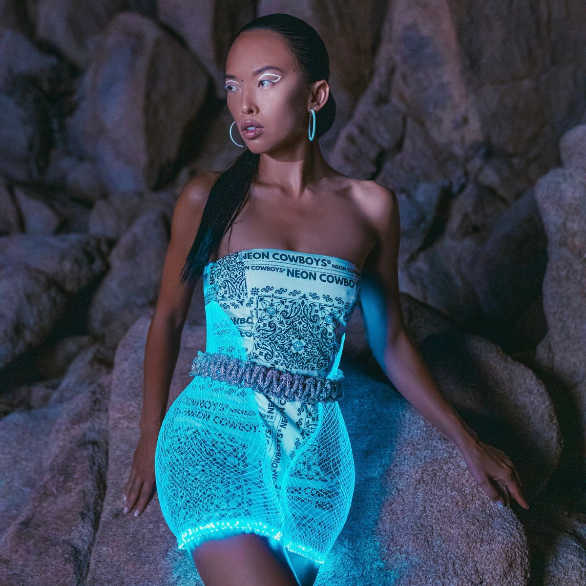Woman wearing blue light up mini bandana net dress against rock backdrop creating stunning glow effect for festival fashion and nighttime events