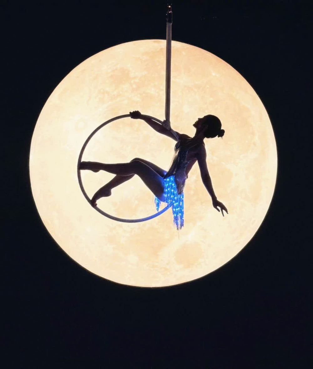Aerial lyra performer in blue Neon Cowboys LED fringe silhouetted against the moon