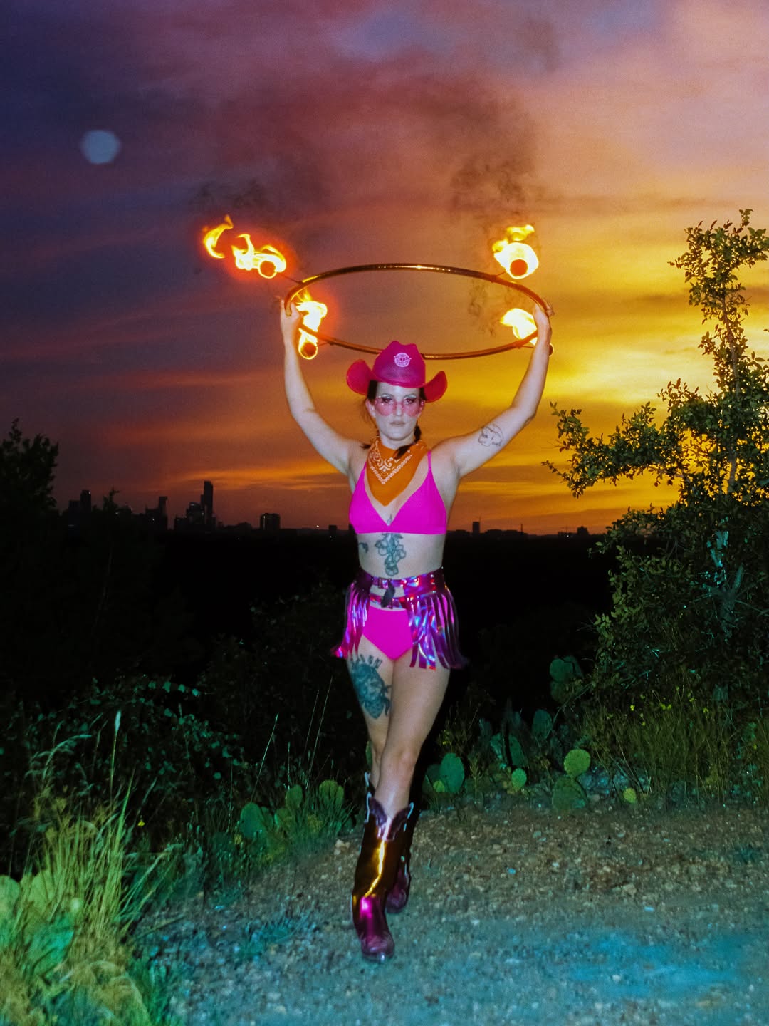 Fire performer in Neon Cowboys pink LED cowboy hat, holographic fringe skirt and chrome boots spinning fire at sunset in Austin Texas, LED and light-up stagewear for performers