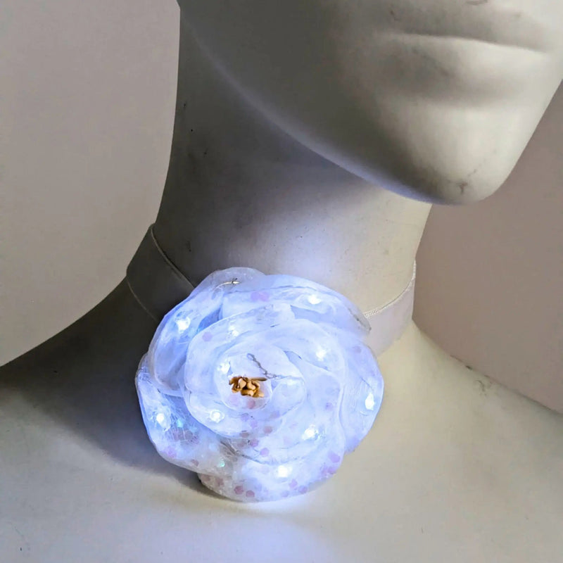 White light up LED flower velvet choker with sequin beaded detail, glowing rose on a soft white fabric band shown close-up on a mannequin, an elegant light up necklace and rave jewelry led necklace for weddings, bachelorette parties, dance performances and night events in the city, Neon Cowboys