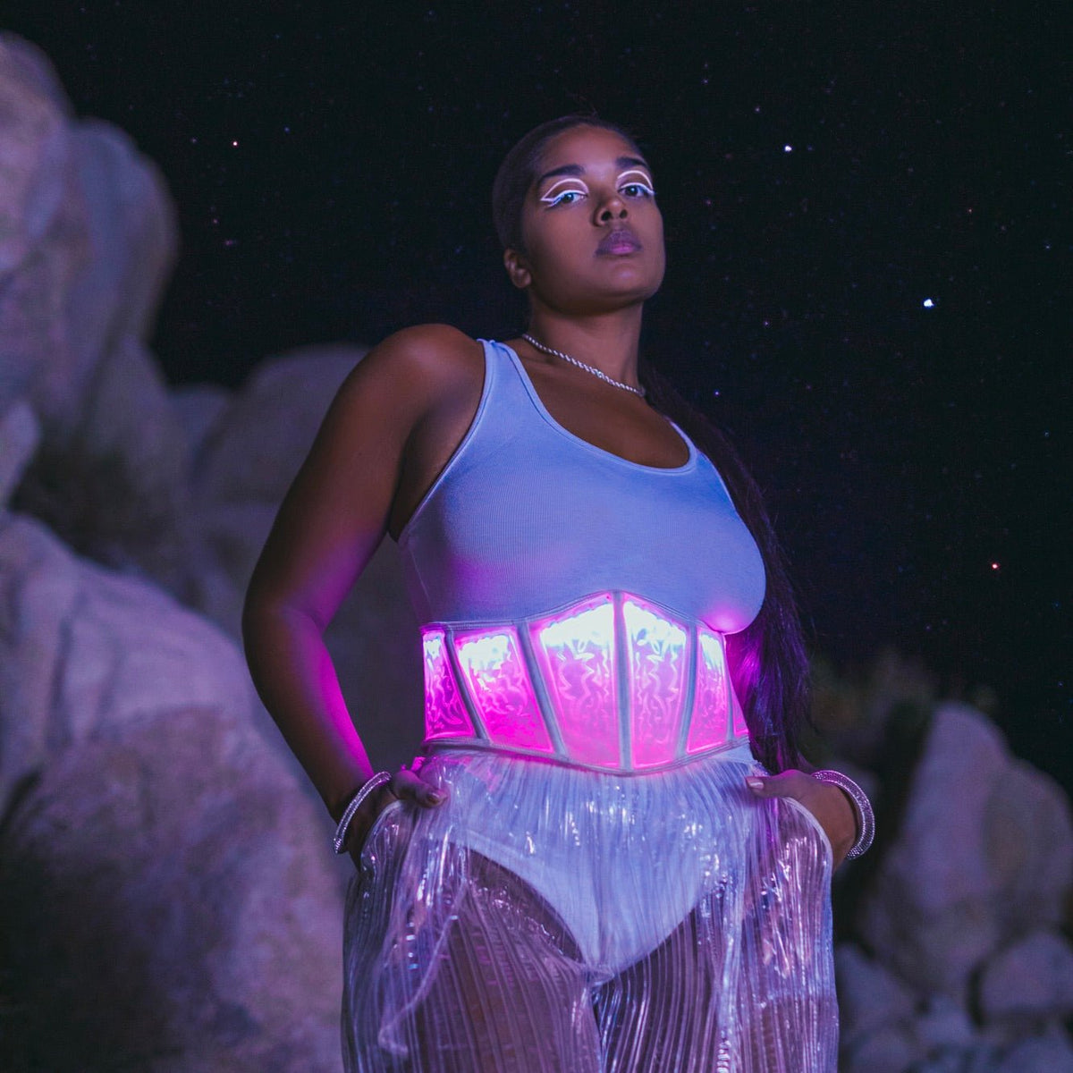 Woman wearing pink programmable LED corset with light up pattern glowing against white outfit in nighttime desert festival setting, perfect for EDM raves and Burning Man