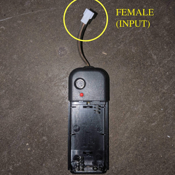 Black battery pack on concrete with a yellow circle highlighting the female input connector, a Neon Cowboys LED replacement inverter pack for light up apparel, hats and led costumes, the spare power source to keep your glow gear running, Neon Cowboys