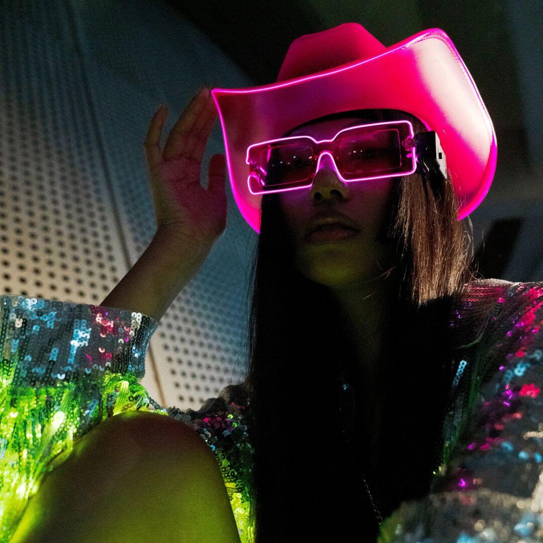 Woman wearing LED neon accessories and glowing pink party sunglasses with LED cowboy hat and light up silver sequin jacket at rave music festival - perfect light up accessories for college, concerts, EDM events, and nightlife parties