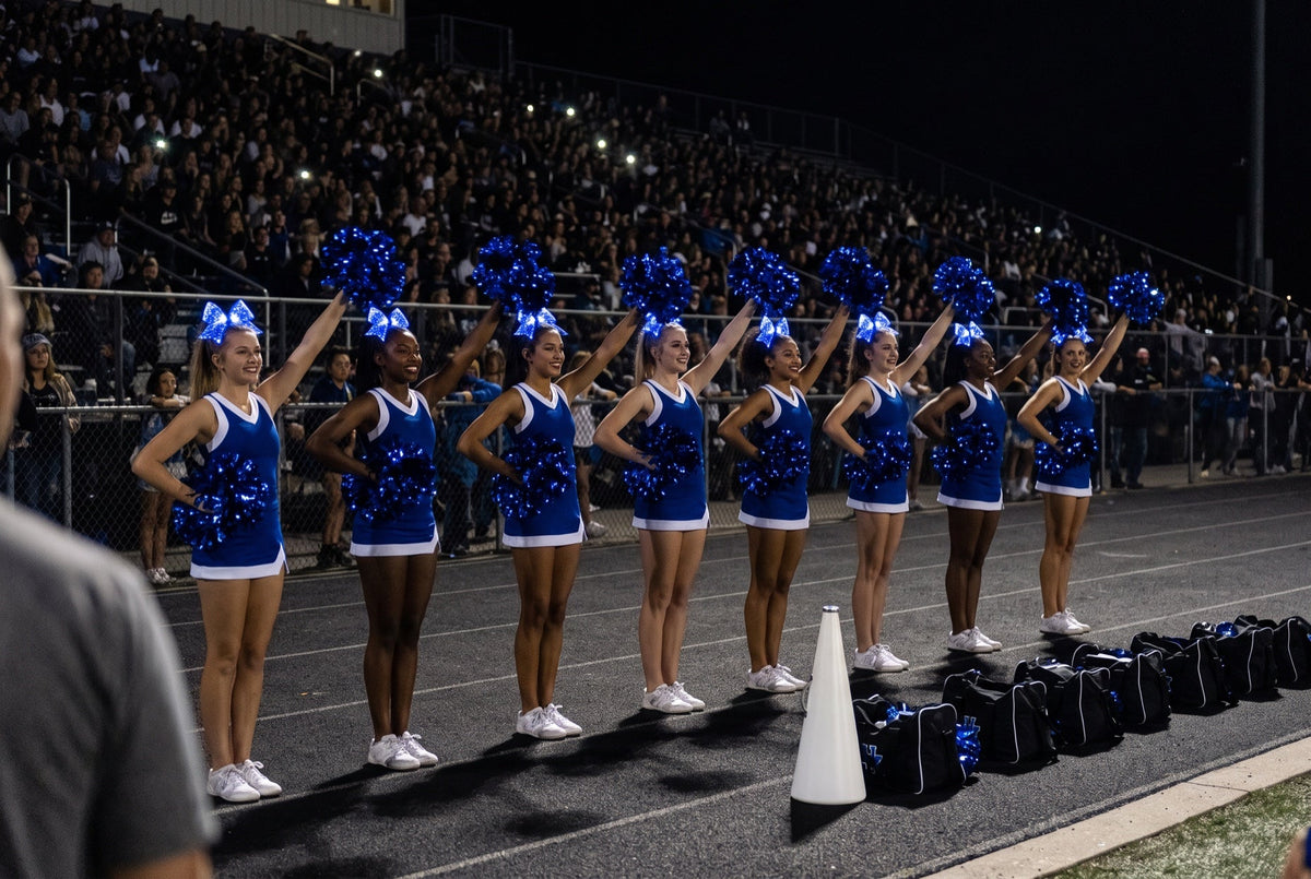 Cheer accessories: light-up LED cheer bows and pom poms glowing royal blue on a college squad on the sideline at night