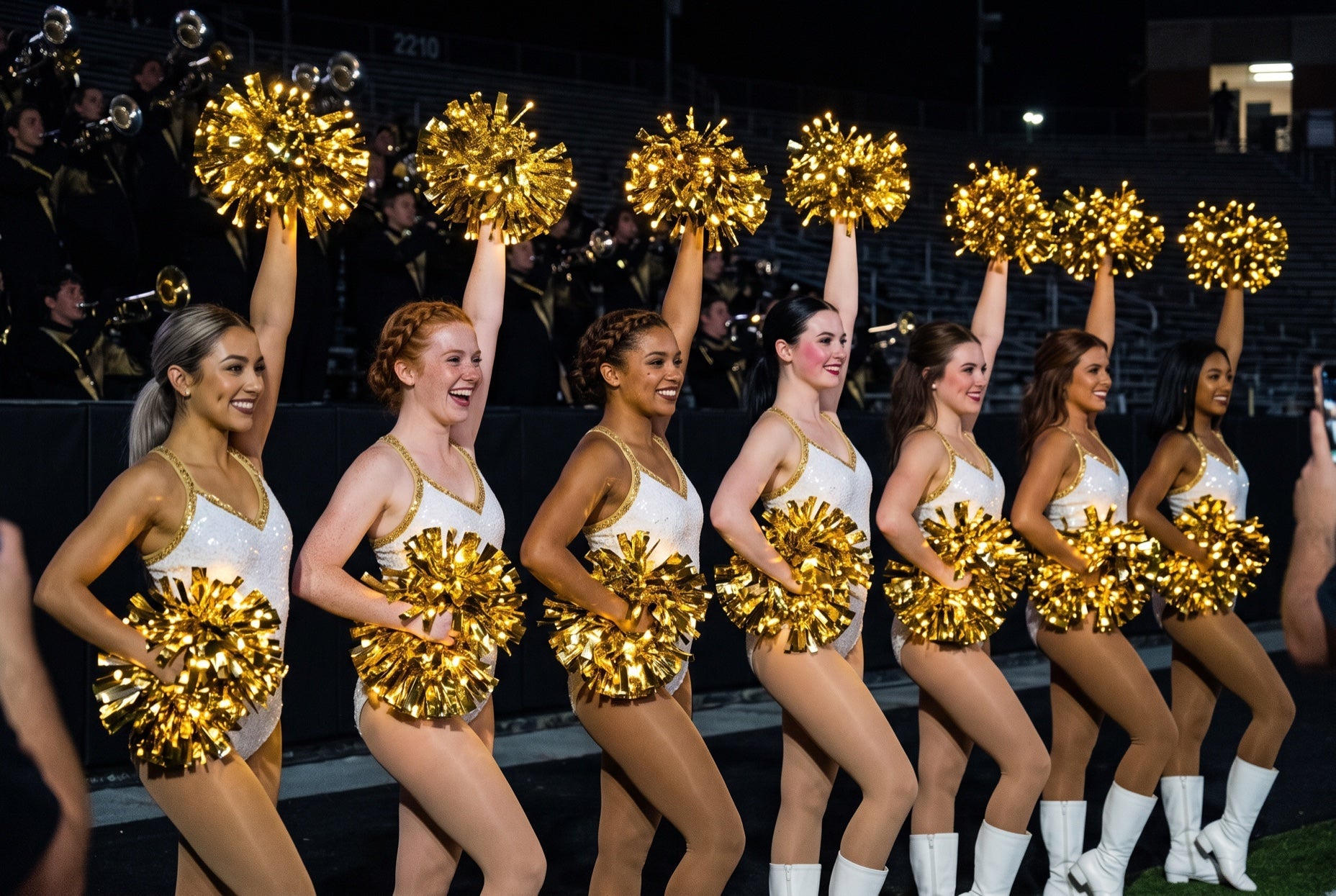 Light up LED gold pom poms glowing on a college band dance line in white and gold with the marching band behind