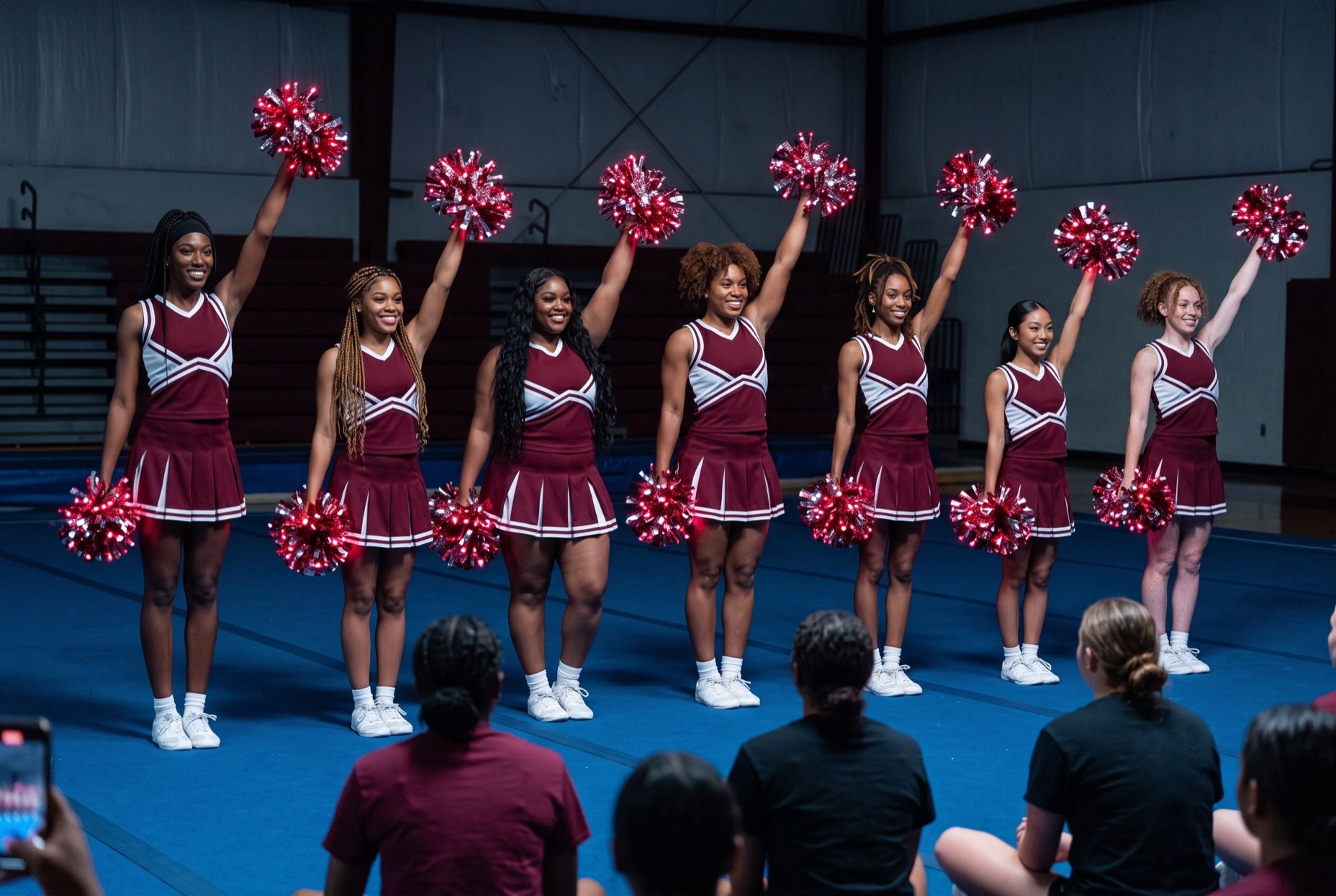 College cheer prep clinic: squad in maroon and white raises crimson LED pom poms on blue mats as adult attendees film