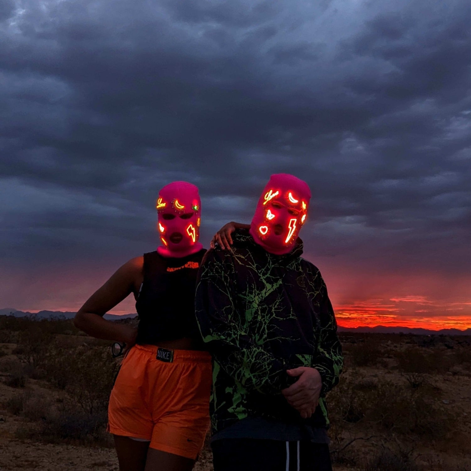 Couple wearing red LED balaclavas at sunset - Matching light-up ski masks for music festivals, desert raves, Burning Man, and outdoor adventure photography