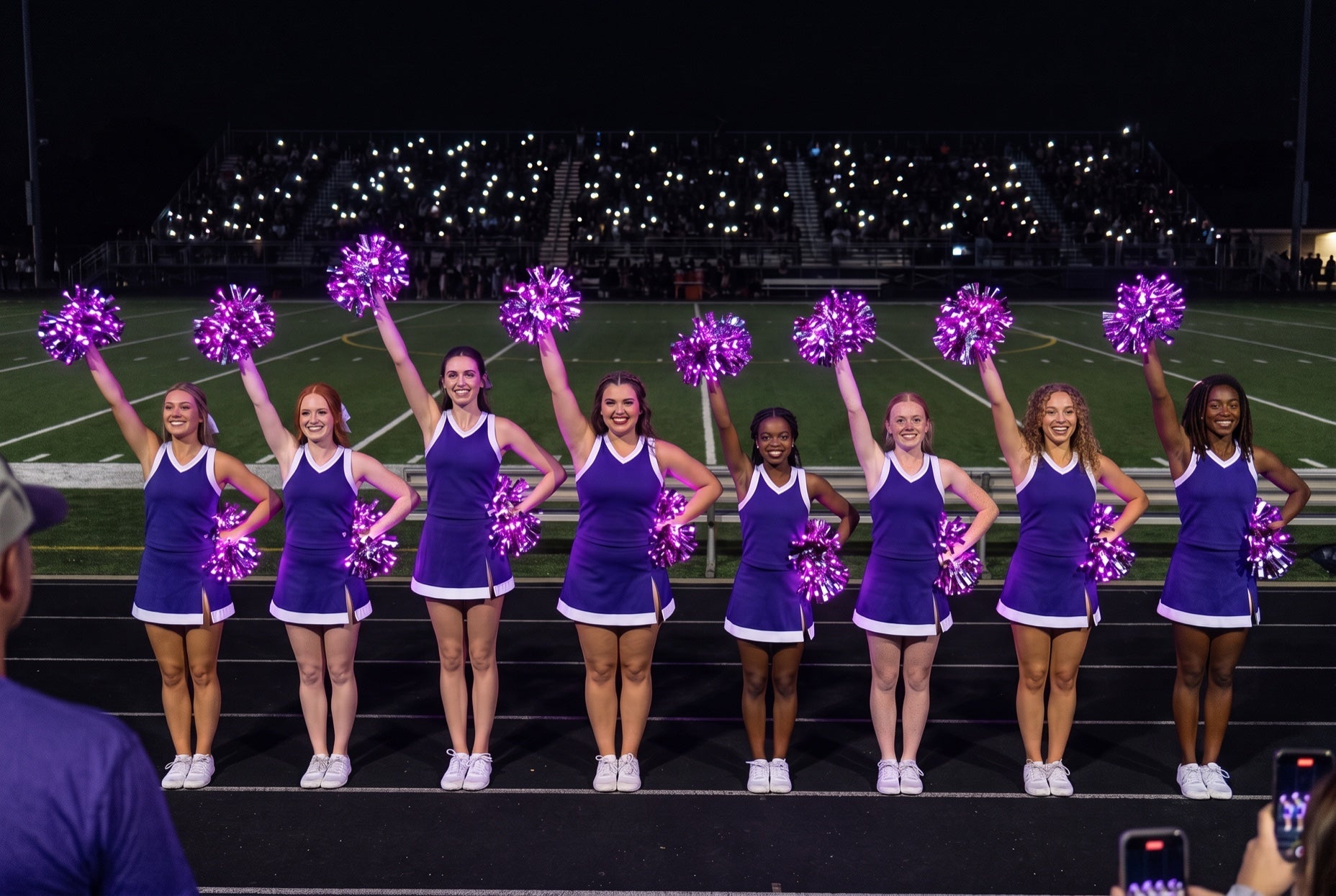 Cheer clinic fundraiser night: college squad in purple raises purple LED pom poms on a dark stadium track at halftime