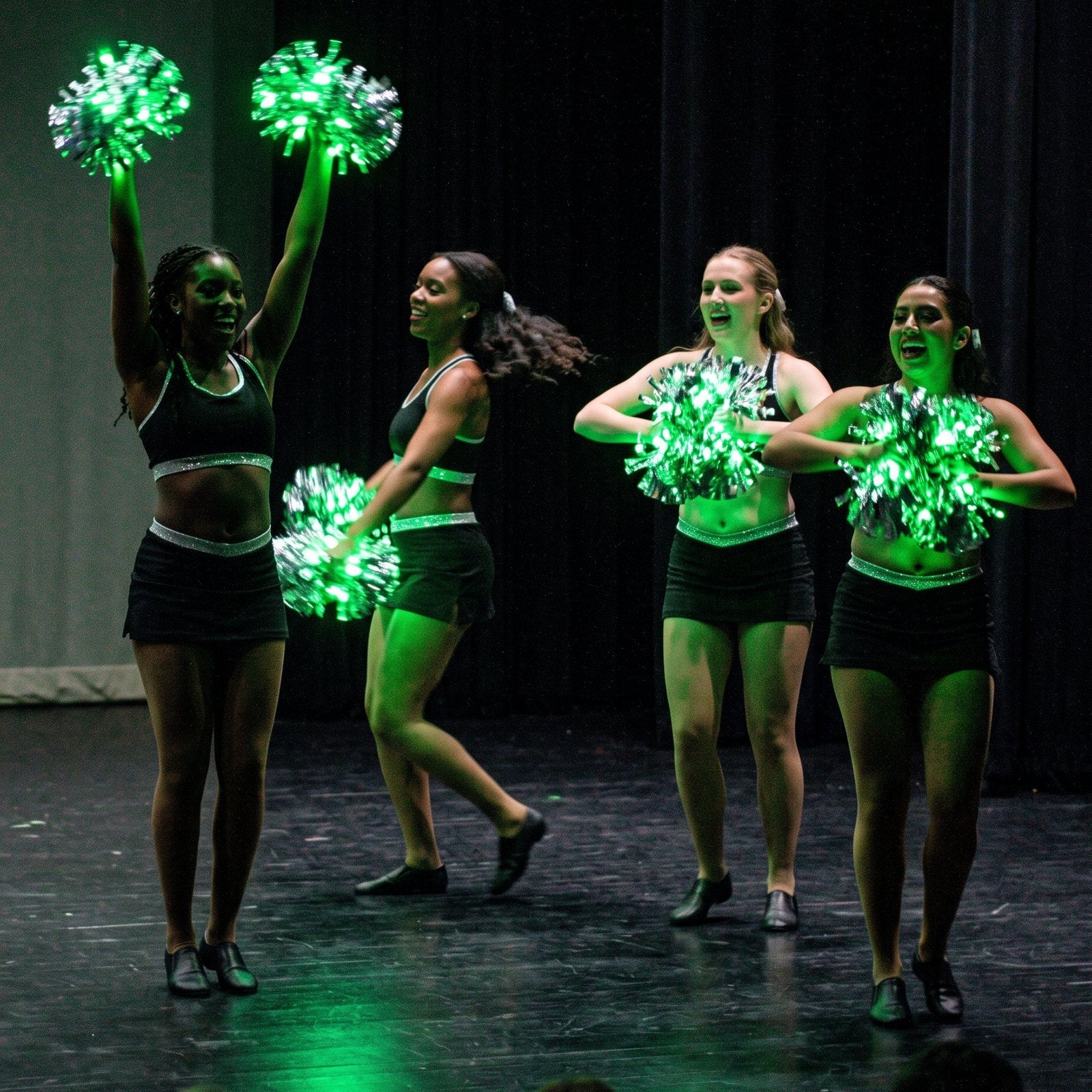 Dance recital on a dark stage with silver LED pom poms glowing green, turning the choreography into a light show