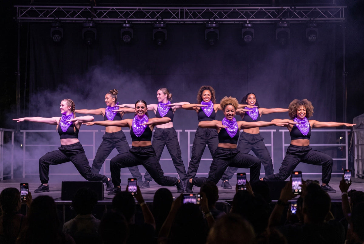 Light up bandanas in purple on an eight-dancer team lunging in unison on an outdoor festival stage at night