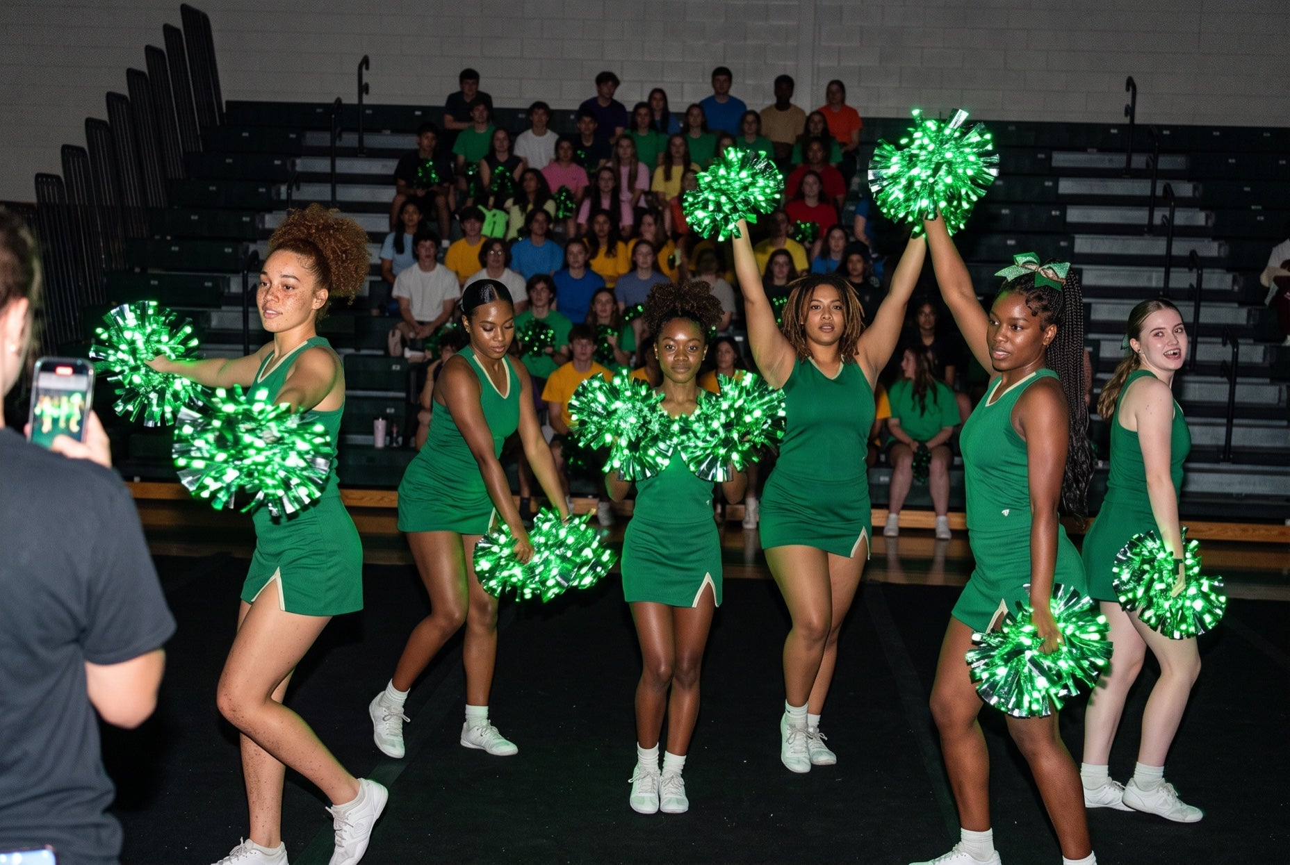 Light up LED pom poms glowing green on a cheer squad leading a spirit week assembly in a dark gym