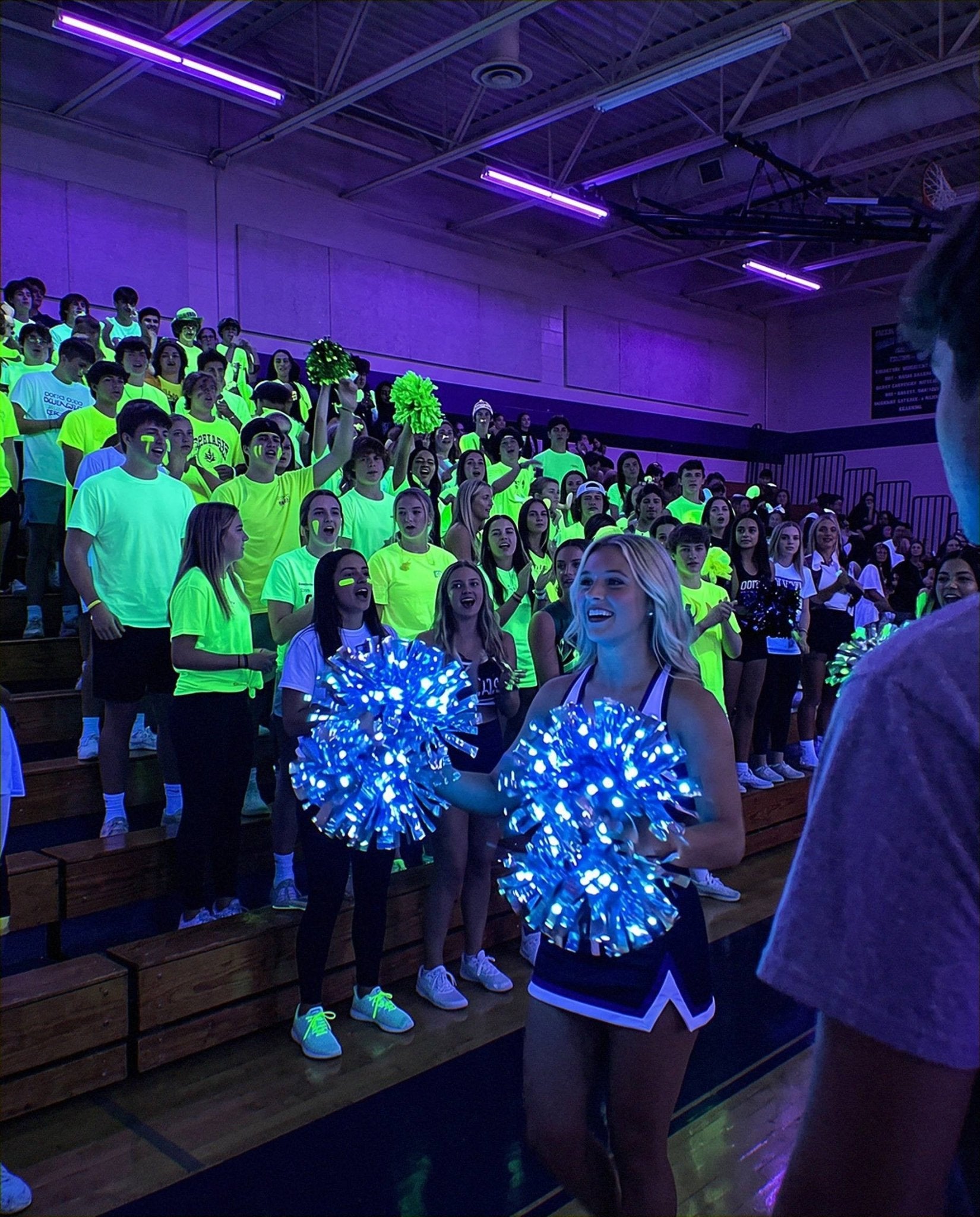 Neon shirts fluorescing green under blacklight at an indoor pep rally while a cheerleader holds LED pom poms glowing blue from their own light.