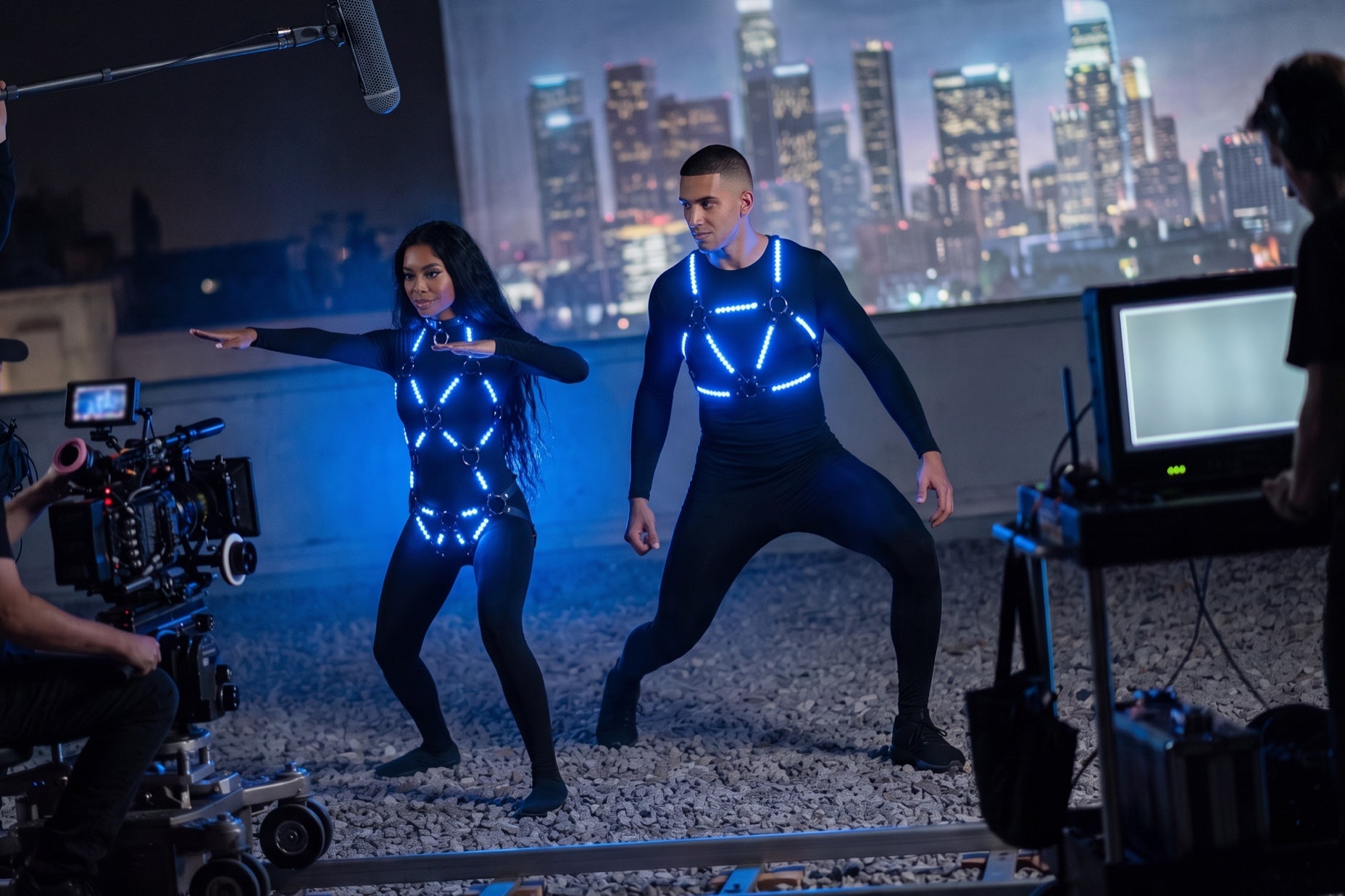Superhero film light-up suit test: two actors in blue LED harnesses rehearse on a rooftop set at night