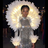 Woman wearing a glowing LED feather aura back piece with matching light-up feather arm cuffs over a silver sequin outfit at an elegant evening event — an illuminated feather halo and elegant light-up accessory for performers, showgirls, galas, festivals, burlesque, circus and stage and event entertainers.