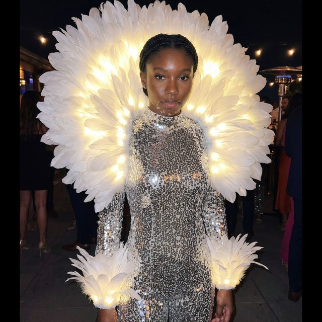 Woman wearing a glowing LED feather aura back piece with matching light-up feather arm cuffs over a silver sequin outfit at an elegant evening event — an illuminated feather halo and elegant light-up accessory for performers, showgirls, galas, festivals, burlesque, circus and stage and event entertainers.