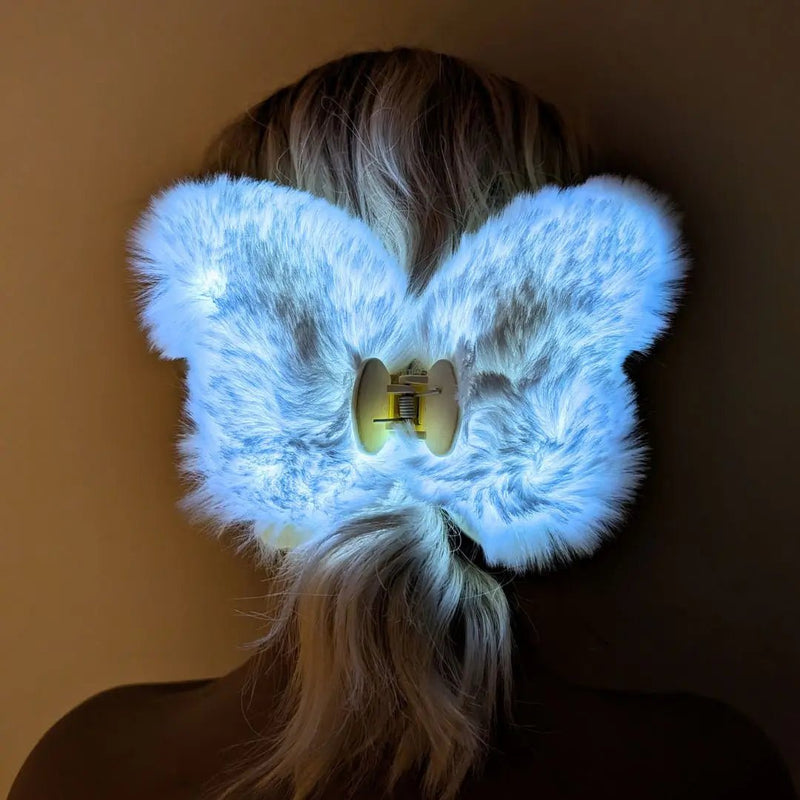 Back of blonde hair wearing a white glowing Neon Cowboys LED fuzzy butterfly claw clip lighting up in a ponytail, a nostalgic 90s inspired light-up hair accessory for Y2K fashion, music festivals, raves and everyday playful hairstyles, Neon Cowboys