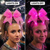 Light-up LED competition cheer bow worn at the crown of a high ponytail, glowing pink, shown with the arena lights on and off