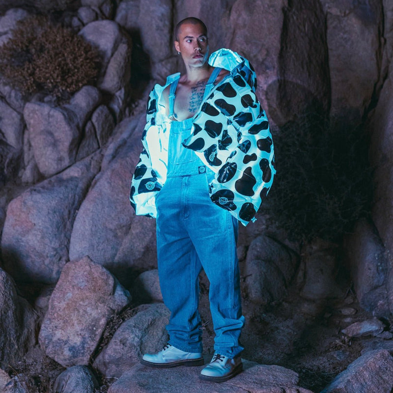 Model wears a Neon Cowboys glowing cow print puffer jacket over blue overalls on desert rocks at night, light up jacket western festival outerwear for raves, EDM concerts, music festivals and country parties, Neon Cowboys