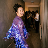 Custom made-to-order LED fringe jacket in purple, a smiling woman at a party in the purple sequin jacket with glowing blue LED fringe, a light up sequin fringe jacket and disco cowgirl jacket in glowing stagewear for go-go dancers, drag and burlesque performers, ideal for music festivals, raves, EDM concerts and Pride, Neon Cowboys