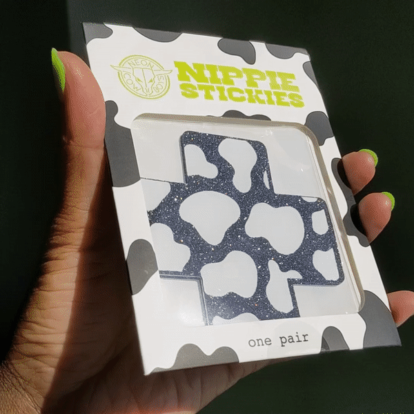 Hand holding open package of X-shaped nipple stickies displaying cow print design perfect for country concerts and western themed events