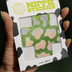 Hand holding open package of cross-shaped nipple stickies showing holographic cow print design that reflects light at concerts and events