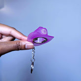 Hand holding purple cowboy hat keychain bottle opener demonstrating size and usability at concerts and nighttime gatherings
