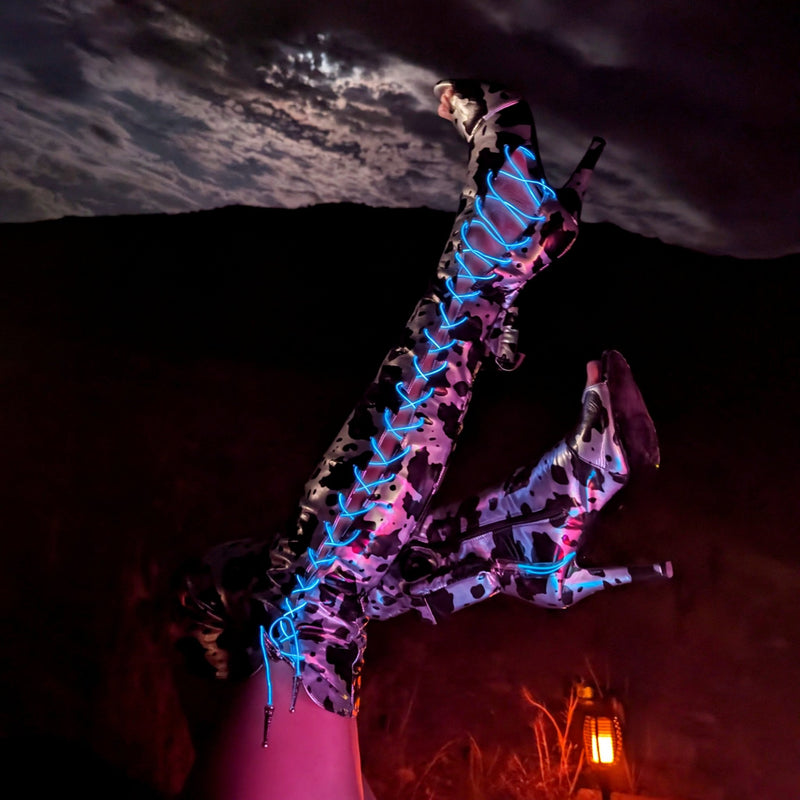 Lace Up Over the Knee Metallic Cow Print Boots, metallic cow print over the knee boots with glowing blue LED laces kicked up against a night sky, light up cowgirl boots and light up heels for space cowgirl and disco cowgirl looks at raves, music festivals, concerts and Halloween, Neon Cowboys