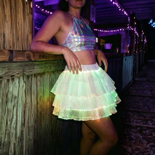 Woman leaning on a wood rail in a green glowing Neon Cowboys LED 3 Layer Tiered Skirt with string lights behind, a light up dress and led rave outfit stagewear for go-go dancers, festival dancers and burlesque performers, a glowing festival outfit for raves, Burning Man and country concerts, Neon Cowboys