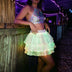 Woman leaning on a wood rail in a green glowing Neon Cowboys LED 3 Layer Tiered Skirt with string lights behind, a light up dress and led rave outfit stagewear for go-go dancers, festival dancers and burlesque performers, a glowing festival outfit for raves, Burning Man and country concerts, Neon Cowboys