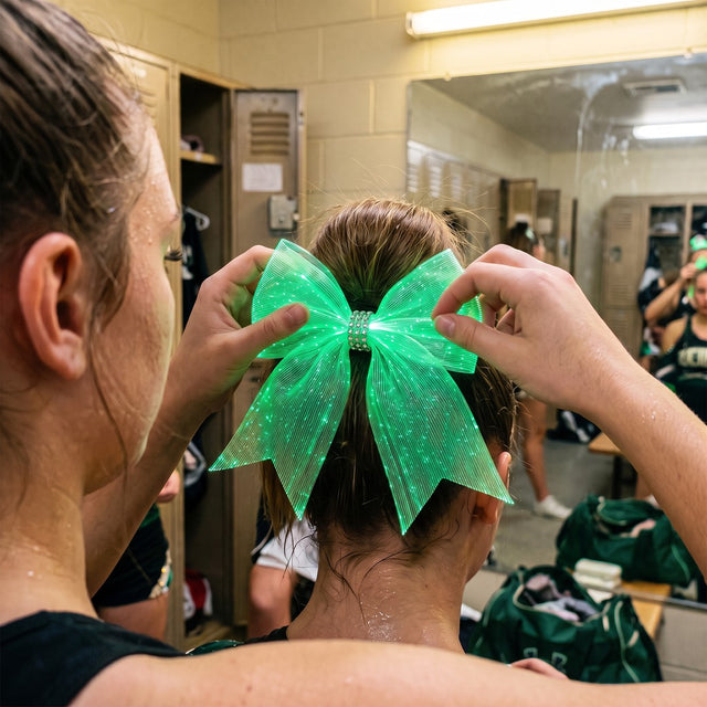 Cheerleader in the locker room tying a light-up LED cheer bow glowing green onto a teammate's high ponytail
