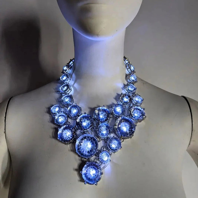 White mannequin bust wearing a chunky clear gem bib statement necklace glowing cool blue, a cool white LED light-up rhinestone jewelry piece with elegant CZ gems, an illuminated led necklace and light up necklace for weddings, formal events, brides and nightclub fashion, Neon Cowboys
