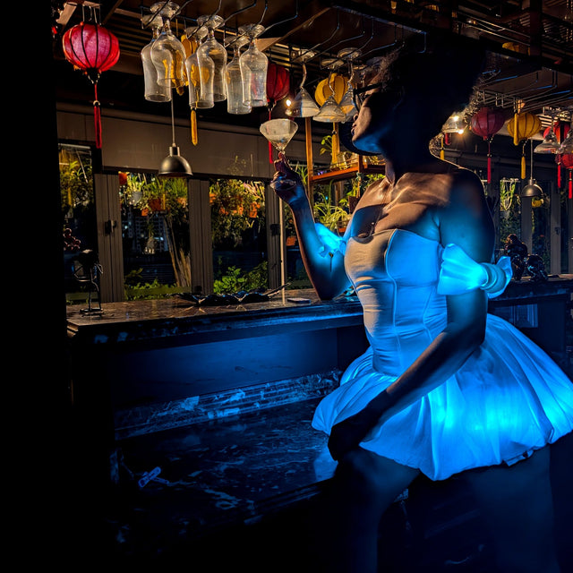Woman sipping a cocktail in a Neon Cowboys LED Fairytale Dream Dress glowing cyan blue at a moody bar, a light up dress and led rave outfit stagewear for showgirls, burlesque performers and drag queens, a dreamy festival outfit for raves, EDM concerts and weddings, Neon Cowboys