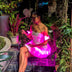 Woman seated in a Neon Cowboys LED Fairytale Dream Dress glowing hot pink in a lantern-lit tropical garden, a light up dress and led rave outfit stagewear for showgirls, cabaret performers and drag queens, a dreamy festival outfit for raves, weddings and bachelorette parties, Neon Cowboys