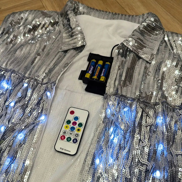 Silver LED sequin fringe jacket laid flat with accessories - Complete package contents showing jacket, remote control, and batteries for color-changing illuminated apparel