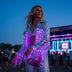 LED Fringe Jacket by Neon Cowboys, a blonde woman at an EDM music festival in a silver light up fringe jacket glowing purple and pink by a neon stage, this led fringe jacket, sequin fringe jacket and disco cowgirl jacket is a rave light up jacket and led costume for festival dancers, showgirls and drag performers at raves, EDC, Coachella and bachelorette parties, Neon Cowboys