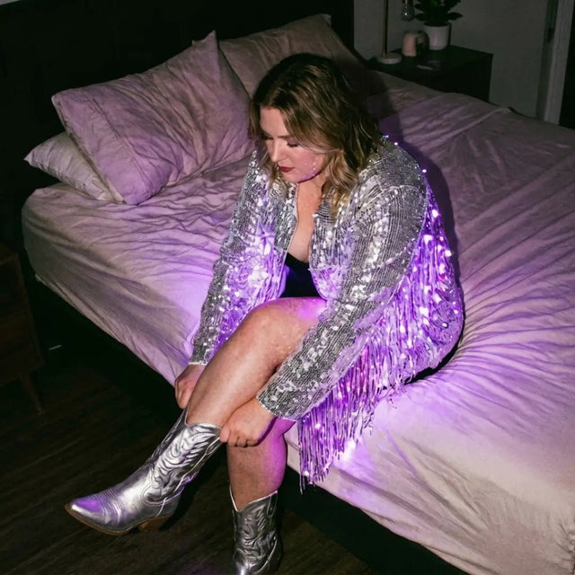 LED Fringe Jacket by Neon Cowboys, a silver light up sequin fringe jacket glowing bright at a night event, this led fringe jacket, sequin fringe jacket and disco cowgirl jacket is a light up jacket and led costume for go-go dancers, showgirls and drag performers at raves, EDM concerts, festivals and bachelorette parties, Neon Cowboys